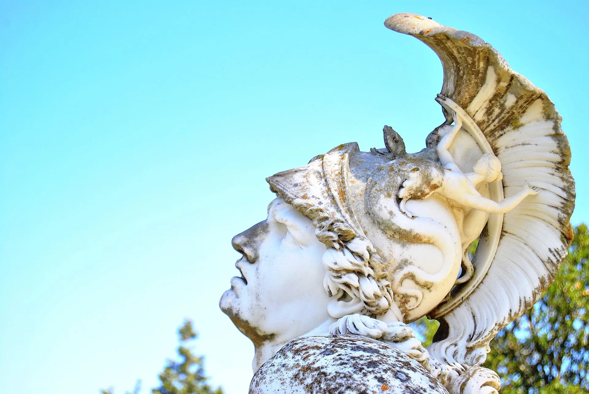 Antike Marmorstatue im Profil, Kopf in den Nacken gelegt, mit muschelartigem Kopfschmuck und Delfin-Relief vor blauem Himmel