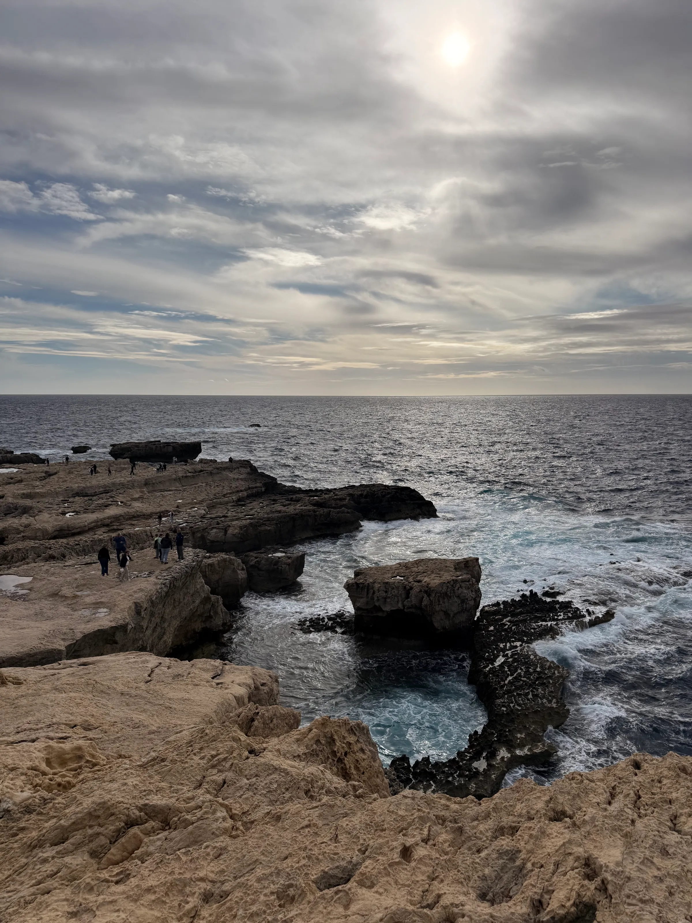 Felsige Kalksteinküste bei Dwejra, Brandung zwischen den Felsplatten, im Hintergrund die Sonne hinter Wolken über dem Meer