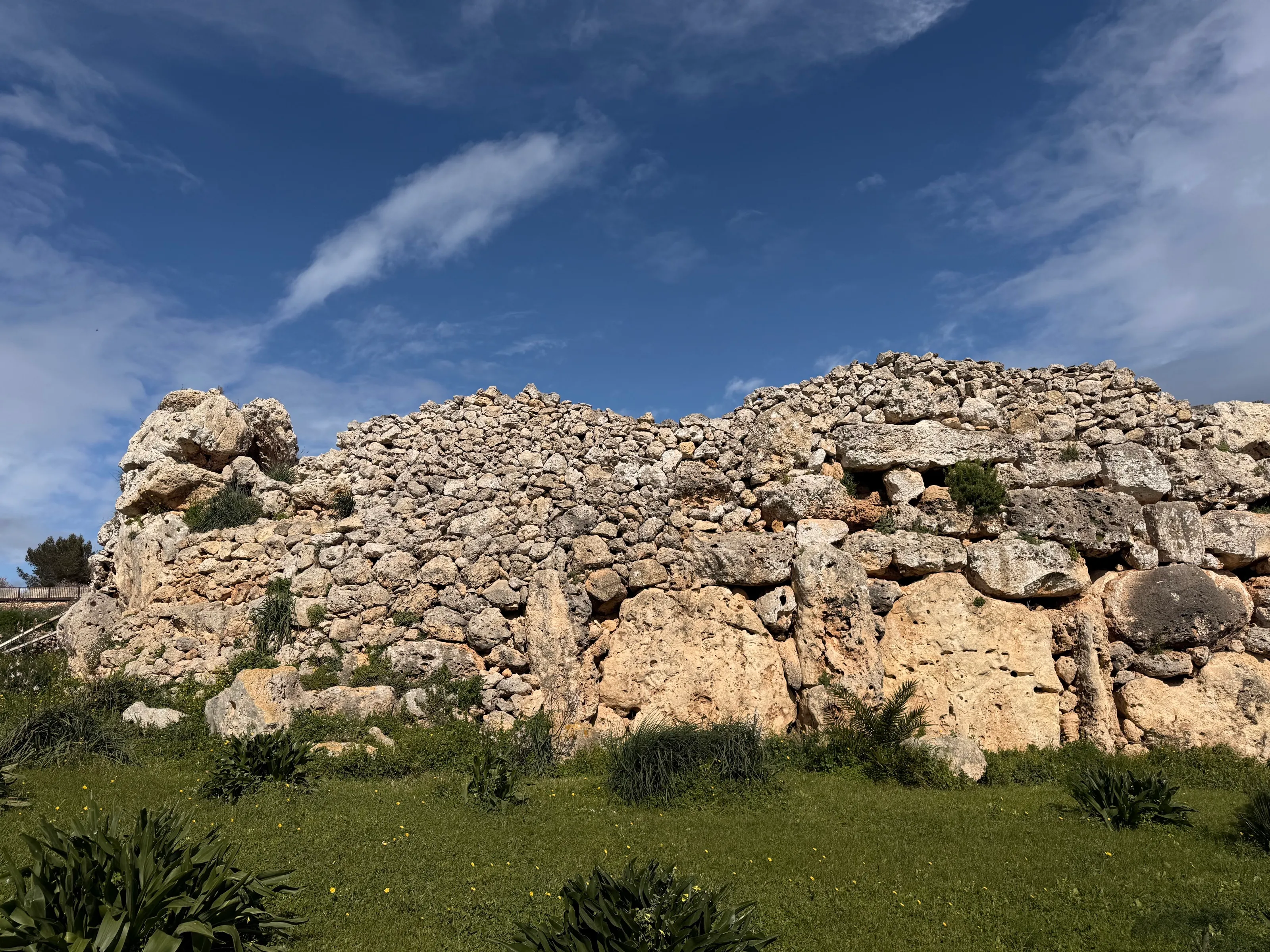 Megalithische Mauer aus großen, unbehauenen Kalksteinblöcken unter blauem Himmel, davor eine Wiese
